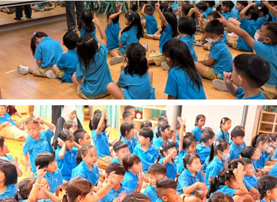 Children in blue uniforms sit on a wooden floor, many raising hands during a classroom activity.